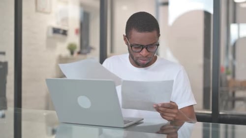 Young Adult Reviews Documents at Laptop in Office