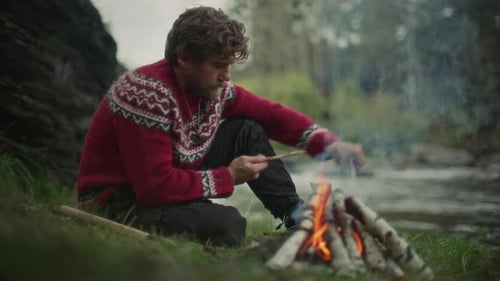 Man Carving Wooden Stick beside Campfire near River in Forest