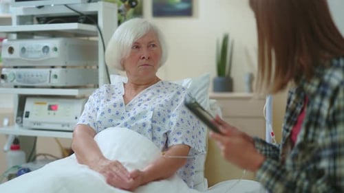 Young Woman Daughter Talking with Her Elderly Mother Patient in Hospital Room Communicating Using