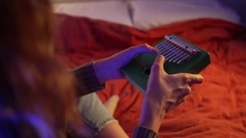 Female Musician Softly Playing Green Kalimba While Resting on Cozy Bed Surrounded By Warm Orange