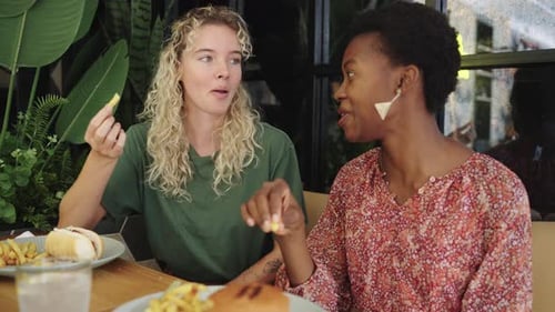 Two Cheerful Multiethnic Women Eating French Fries in a Restaurant