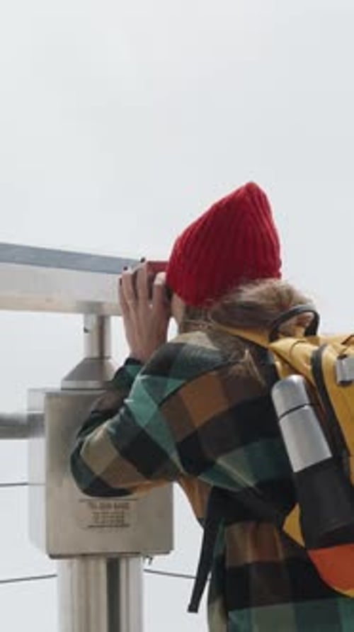 Vertical video. A female tourist looks through binoculars at a high mountain peak in the clouds.