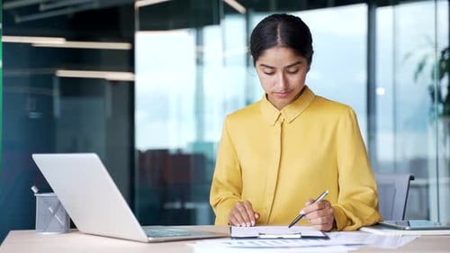 Confident young businesswoman sitting at desk in modern office checking and signing documents.