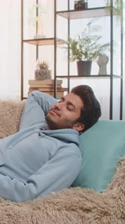 Tired Indian Man Lying on Comfortable Couch Taking Rest and Daytime Nap in Living Room at Home