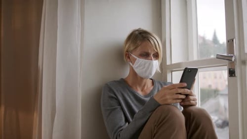 Worried woman with face mask using smartphone indoors