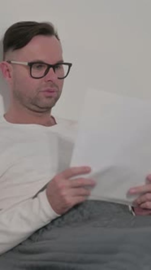 Man Reads Documents in Bed Under Blanket