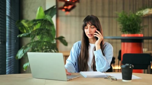 Happy Businesswoman Engaged in Phone Call While Working on Laptop in Modern Office