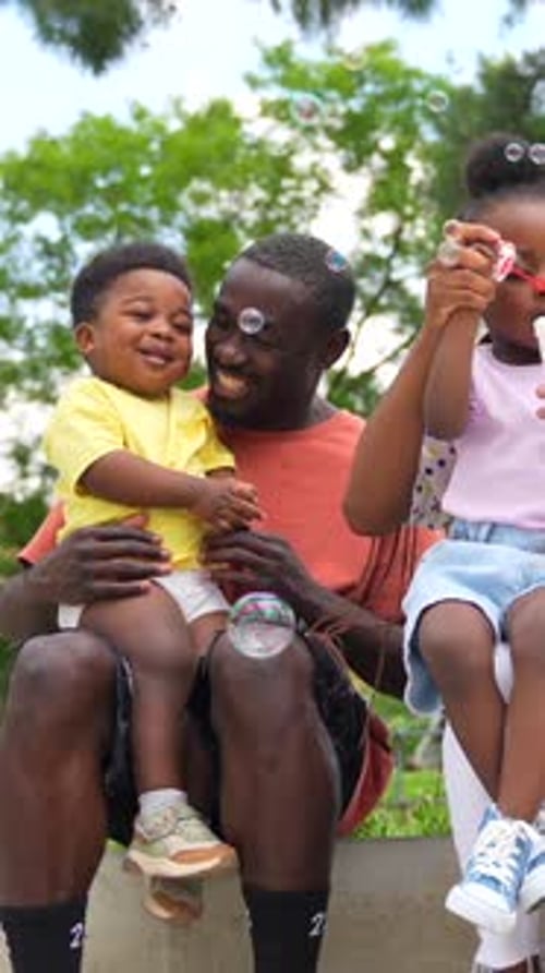 African black ethnicity family with children in playground having fun blowing soap bubbles next to t