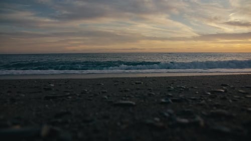 Wide view of wave breaking on pebbled Spanish beach during pastel sunset by the sea