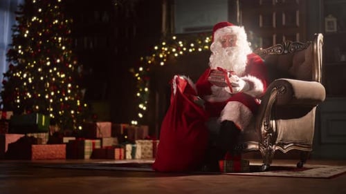 Santa Claus Filling Christmas Bag by Lit Tree
