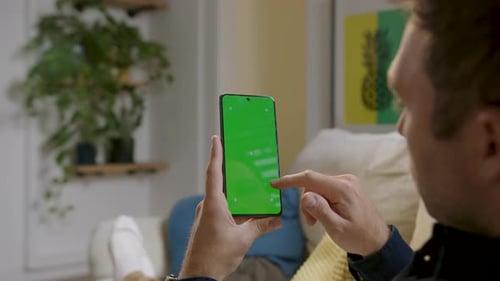 Man comfortably seated at home, holding a smartphone with green screen and interacting with it as if