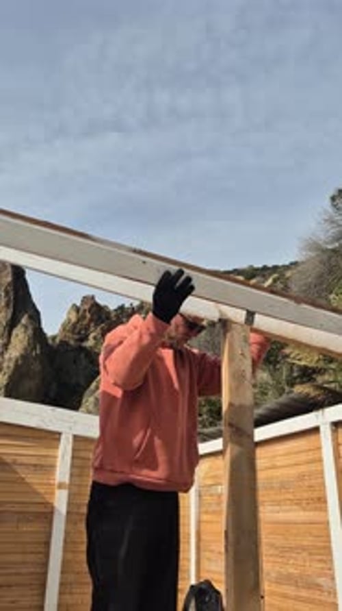 Construction Worker Dismantling Wood Plank Framework Outdoors on Sunny Day Near Rocky Hills