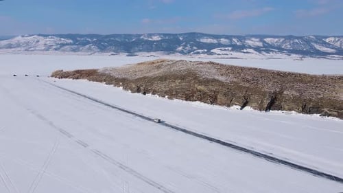 Winter Road Cuts Through Snowy Mountains Passing By a Frozen Lake Seen From an Aerial View