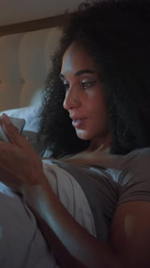 Une femme afro-américaine accro aux gadgets gagne avec un téléphone portable allongée dans son lit à Dark Night Happy