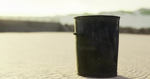 Black Trash Bin Stands Alone on Sandy Beach Under Bright Sunlight