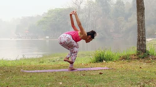 Asian woman practicing yoga doing Reverse Eagle Pose on the mat beside a lake in outdoor park.