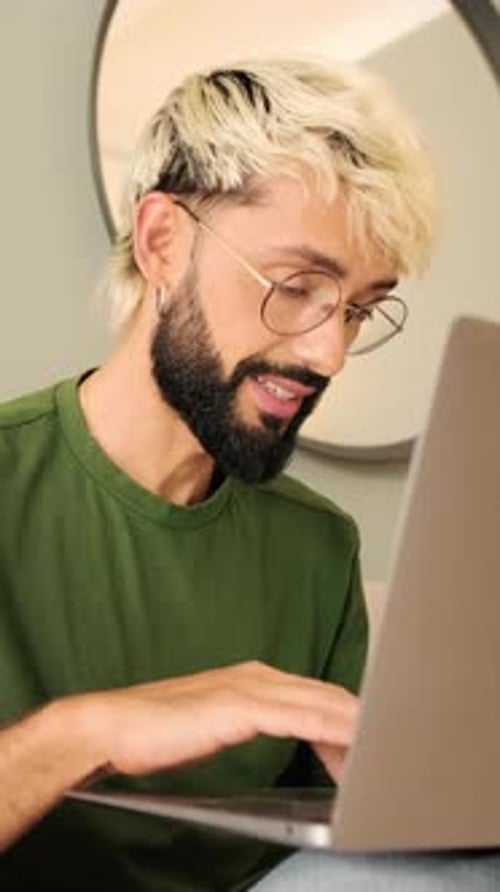 Young blond man with a beard works on a laptop while sitting in a cozy living room