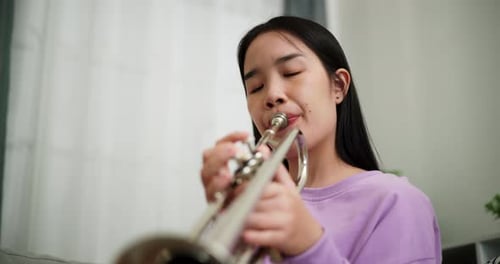 A young woman practices playing the trumpet while sitting on a sofa in a cozy living room.