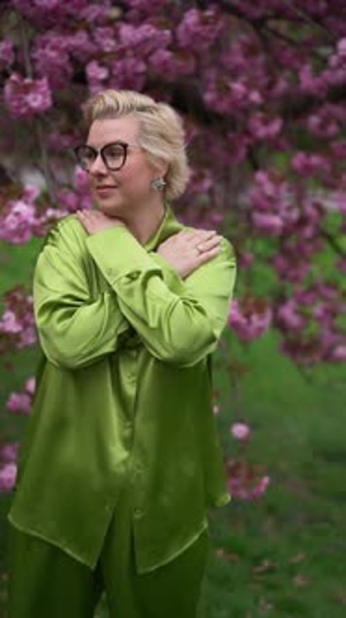 Woman in Green Silk Shirt Poses Amid Blooming Pink Blossoms Outdoors