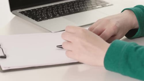 Hands Write on Clipboard Next to Laptop