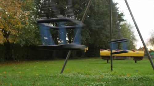 A side on STATIC SHOT of two empty swings swaying fast with no people around, in a desolate playgrou
