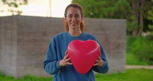 Smiling Woman Offers Red Heart Balloon in Park