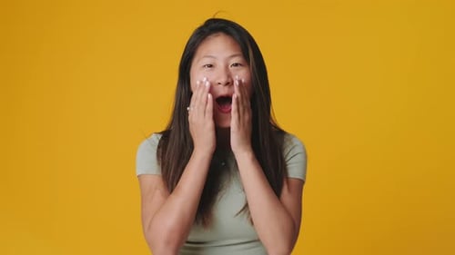 Young woman raising hands in surprise looking at camera, isolated over yellow background in studio