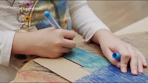 Girl Paints Cardboard Figure with Blue Marker at Table