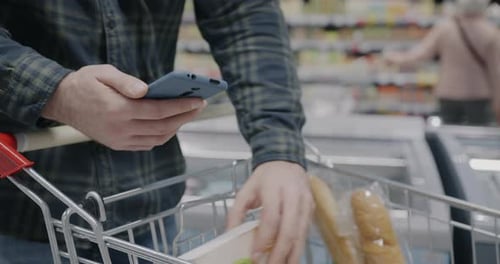 Closeup of Male Hand Using Smartphone While Man Holding Shopping Trolley in Supermarket