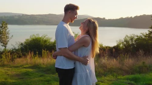 Young Couple in White Clothes Hugging and Cuddling in the Fields with Lake View at Sunset