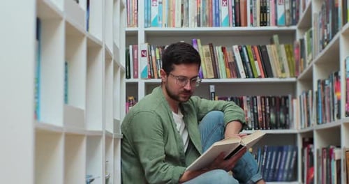 Caucasian Student Man Reading Book Sitting Among Stack of Books on Library Floor Side View
