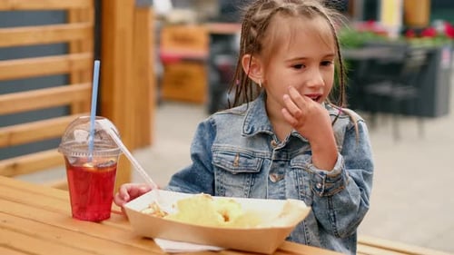 A girl with pigtails eats fast food with her hands, licking her fingers and enjoying lunch in a cafe