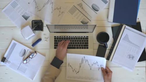 Top View Arms of Young Businessman Looking at Diagrams with Statistics on Papers and Typing Text on