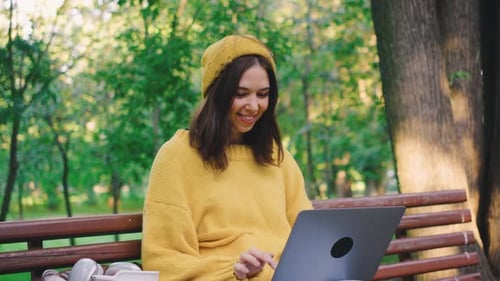 A Cheerful Woman is Working on Her Laptop While Seated Comfortably on a Bench in the Park Fully
