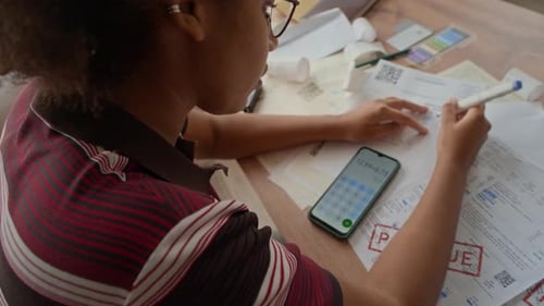 Woman Reviewing Overdue Bills and Using Phone Calculator While Managing Finances
