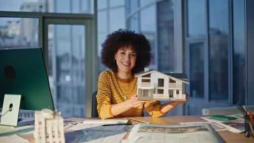 Ingeniero profesional mostrando un modelo de casa a la cámara en la oficina, una mujer sonriente