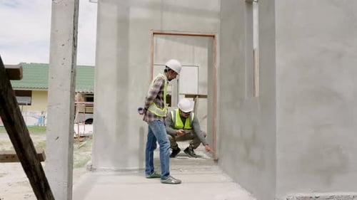 Construction engineer and Asian foreman examine cement wall and construction plans from blueprints.