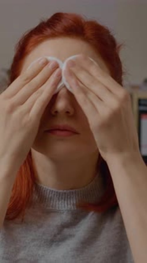 Vertical video Young woman with red hair removes her makeup in front of the mirror at home