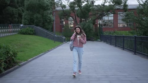 Young Woman with a Backpack and Coffee Walks Along a Paved Outdoor Path While Talking on Her Phone