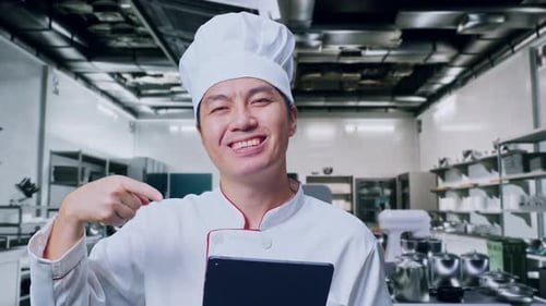 Close Up Of Asian Man Chef Smiling And Pointing To Tablet In His Hand In Kitchen