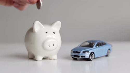 Hand putting coin into piggy bank next to toy car. Piggy bank with car on a wooden table.