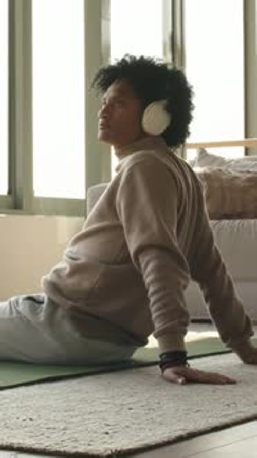 Vídeo vertical: Hombre inclinado con suéter y auriculares que se mueve hacia atrás sobre la alfombra junto a la ventana para escuchar audio guiado