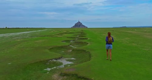 A Female with a Backpack Visits the Mont Saint Michel Castle While Walking Through Green Meadows