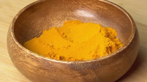 Spoon picks up Turmeric Powder spice from wooden bowl and rotating on wooden board. Dry seasoning