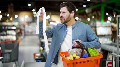 Unhappy customer holding a long grocery receipt check. Man feeling shocked about rising grocery