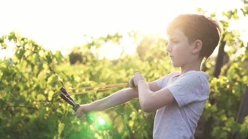 Concentrated boy shooting wooden slingshot a pebble at a target against of a green vineyard