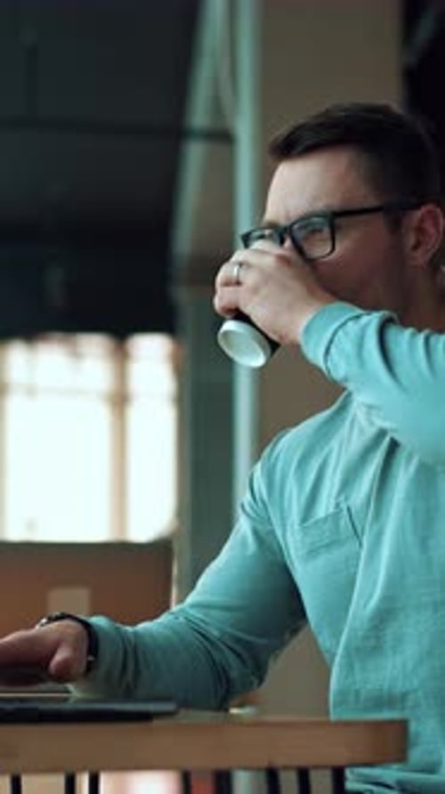 Man working on laptop and drinking coffee from a paper cup.