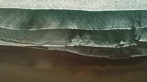Aerial view of breaking ocean waves rolling onto a dark sandy tropical beach