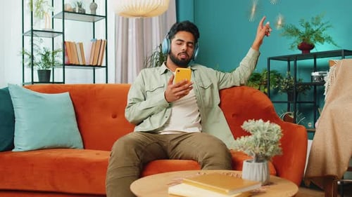 Un homme heureux et détendu dans des écouteurs sans fil danse en écoutant de la musique rock n roll préférée sur son canapé