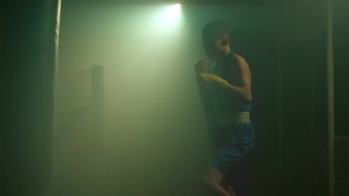 Boxeador masculino en forma en ropa deportiva entrenando sobre fondo ahumado con luz